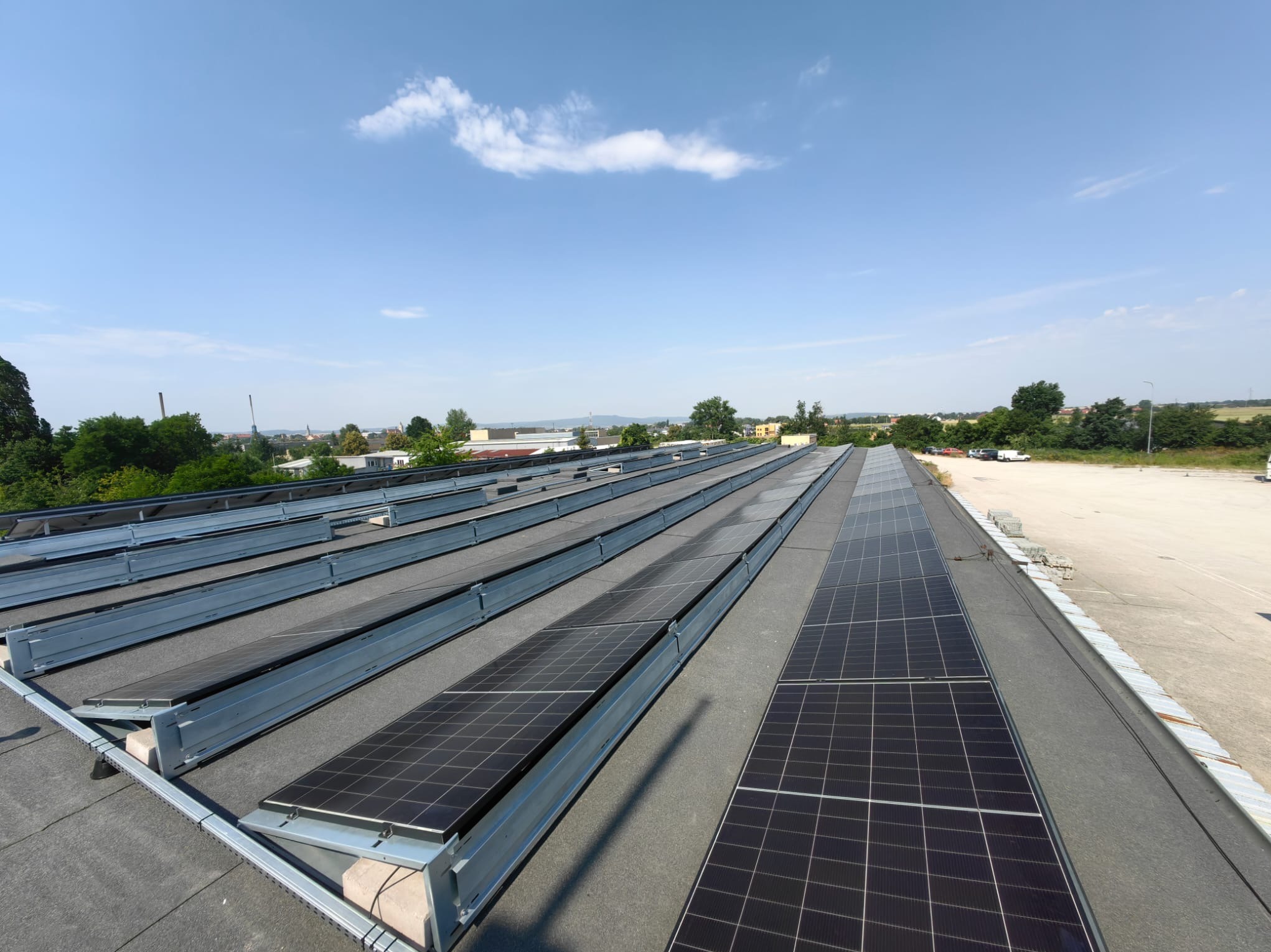Instalacja fotowoltaiczna na dachu budynku, panele w metalowych ramach, widok z góry, błękitne niebo z chmurami w tle, teren zabudowany w oddali.