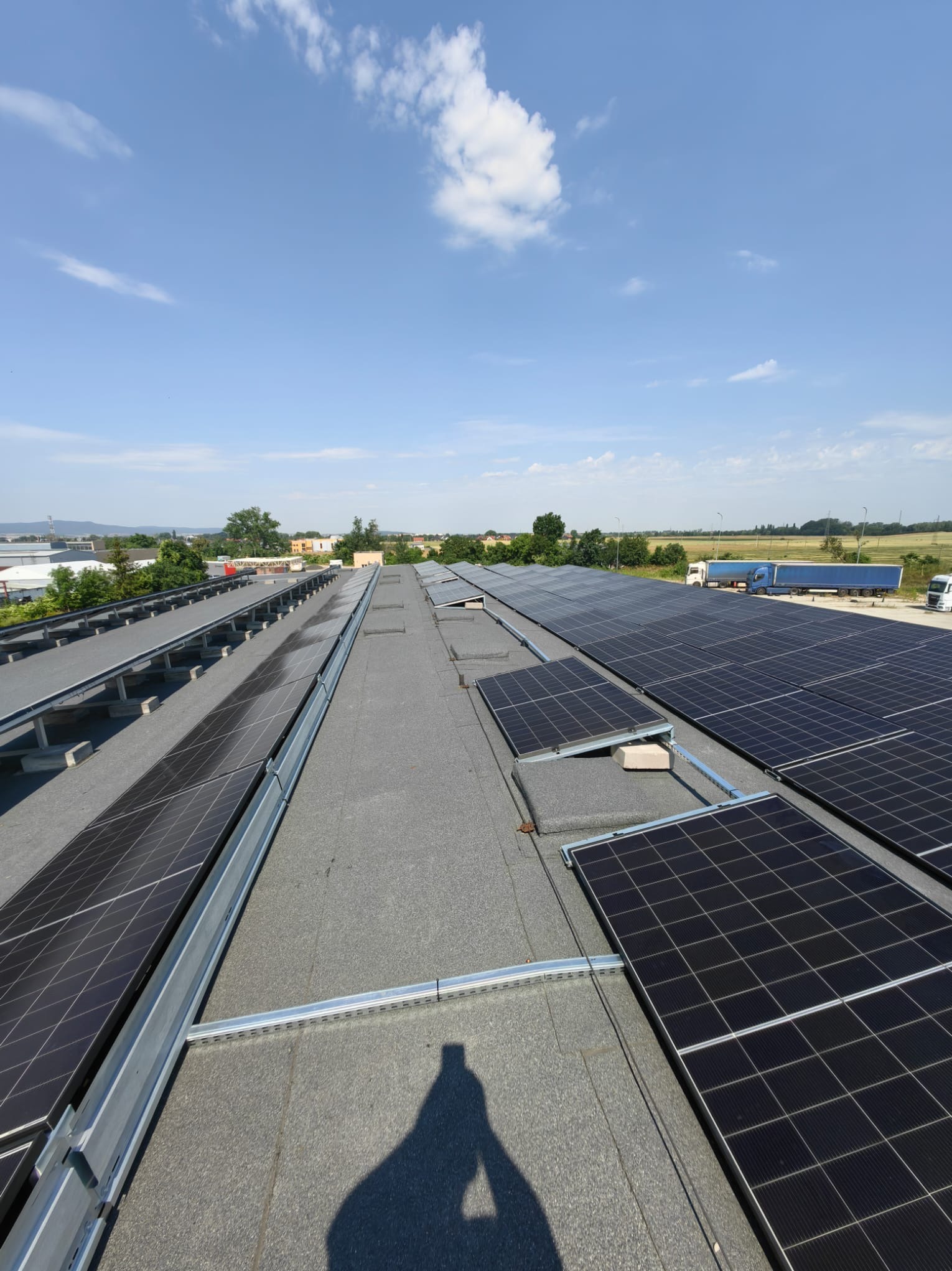 Instalacja fotowoltaiczna na dachu budynku przemysłowego w słoneczny dzień. Widoczne panele, system montażowy oraz cień osoby wykonującej zdjęcie. W tle zabudowania i niebo.