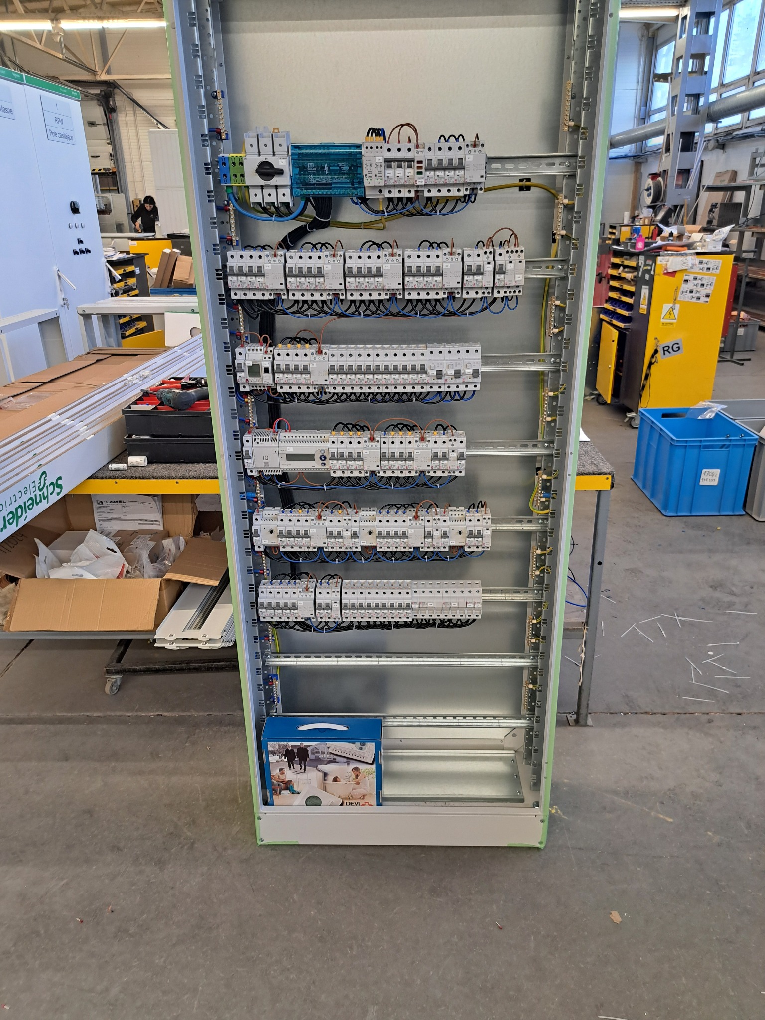 Rozdzielnica elektryczna z zainstalowanymi bezpiecznikami i okablowaniem, widoczna w hali produkcyjnej, z pudełkiem akcesoriów DEVI u dołu.