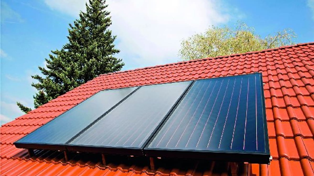 Czerwony dach z trzema panelami solarnymi w czarnych ramach, widoczne drzewo i niebieskie niebo w tle. Instalacja solarna na tle błękitnego nieba.