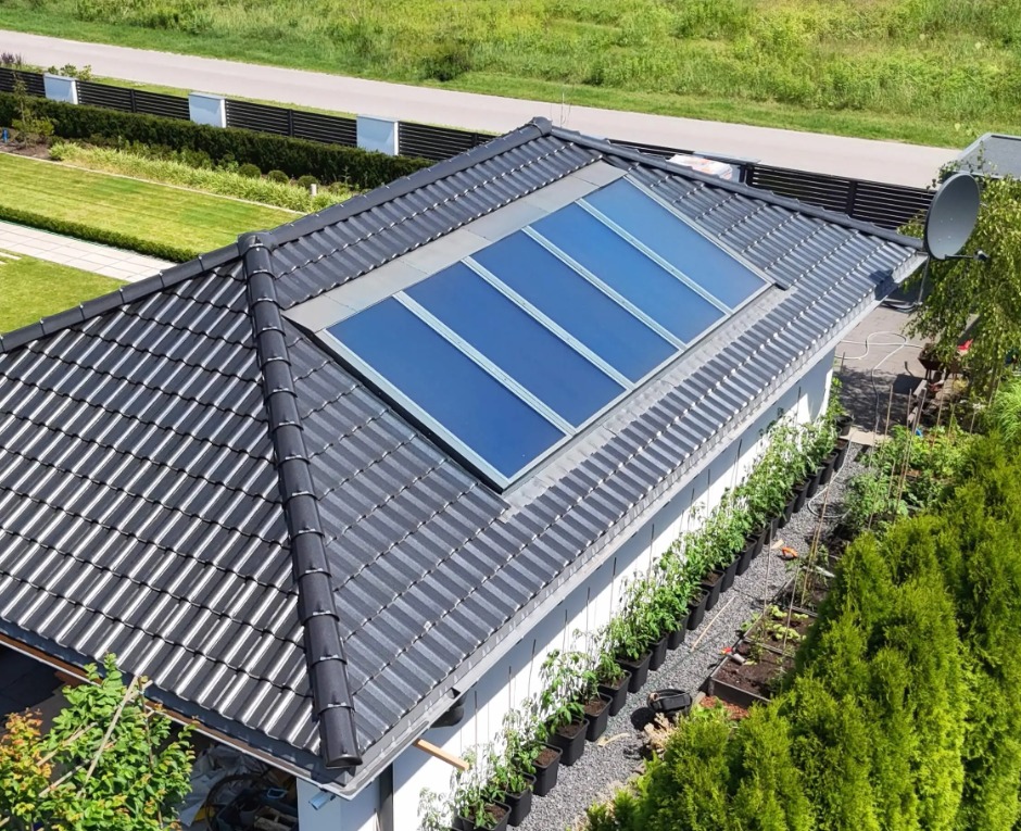 Czarny dach z panelami solarnymi wkomponowanymi w konstrukcję, widok z góry. Obok domu ogród z roślinami w donicach i żywopłotem, w tle droga i zieleń.