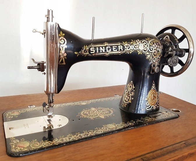 Antyczna maszyna do szycia Singer, czarna z ozdobnymi, złotymi detalami floralnymi, widoczna igła i koło zamachowe, umieszczona na drewnianej podstawie.