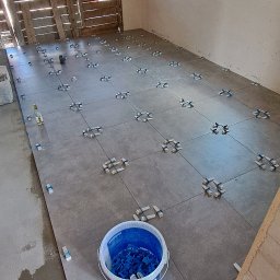 PCH usługi remontowo-budowlane - Układanie szarych płytek podłogowych z użyciem klipsów poziomujących w Nowym Sączu. Widoczny kubeł z klipsami i drzwi wejściowe z drewnianą kratą.