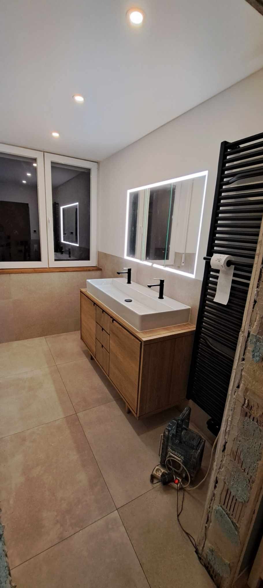 Nowoczesna łazienka z podwójną umywalką nablatową i podświetlanym lustrem. Na podłodze beżowe płytki, widoczny grzejnik i narzędzia.