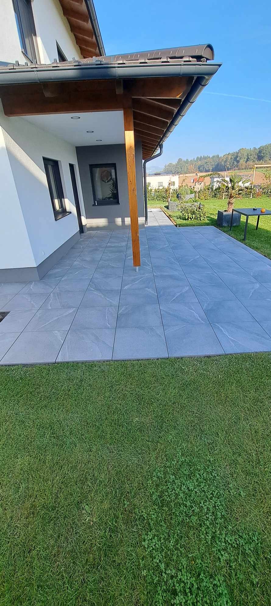 Nowoczesny taras z szarych, kwadratowych płytek, przylegający do domu z drewnianym zadaszeniem i zadbanym trawnikiem. Widok na ogród i zabudowę w tle.