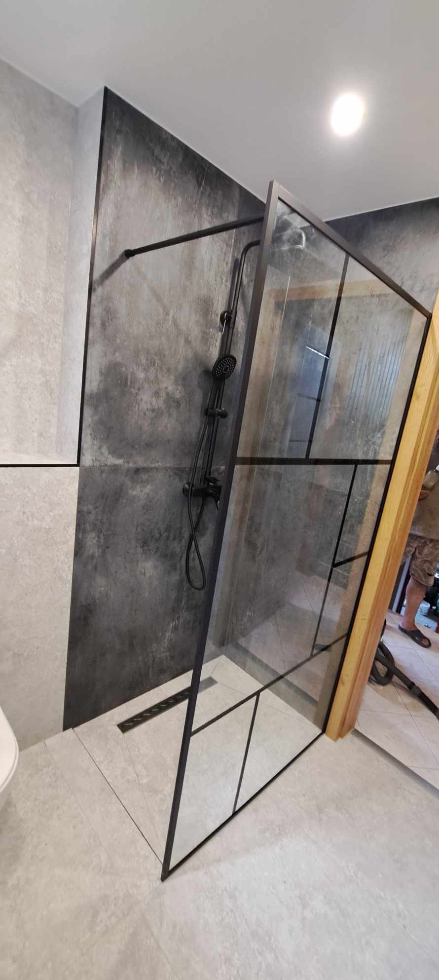 Nowoczesna kabina prysznicowa z czarnymi profilami i deszczownicą na tle szarej ściany w łazience. Widoczny odpływ liniowy i jasne płytki podłogowe.