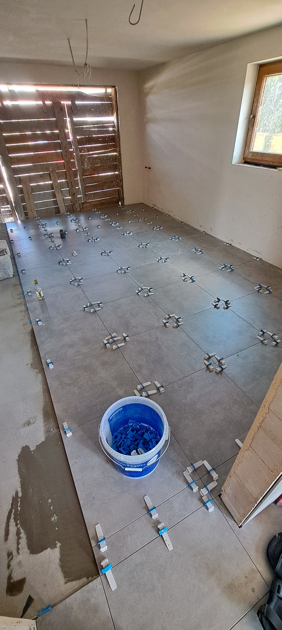 Układanie szarych płytek podłogowych z użyciem klipsów poziomujących w Nowym Sączu. Widoczny kubeł z klipsami i drzwi wejściowe z drewnianą kratą.