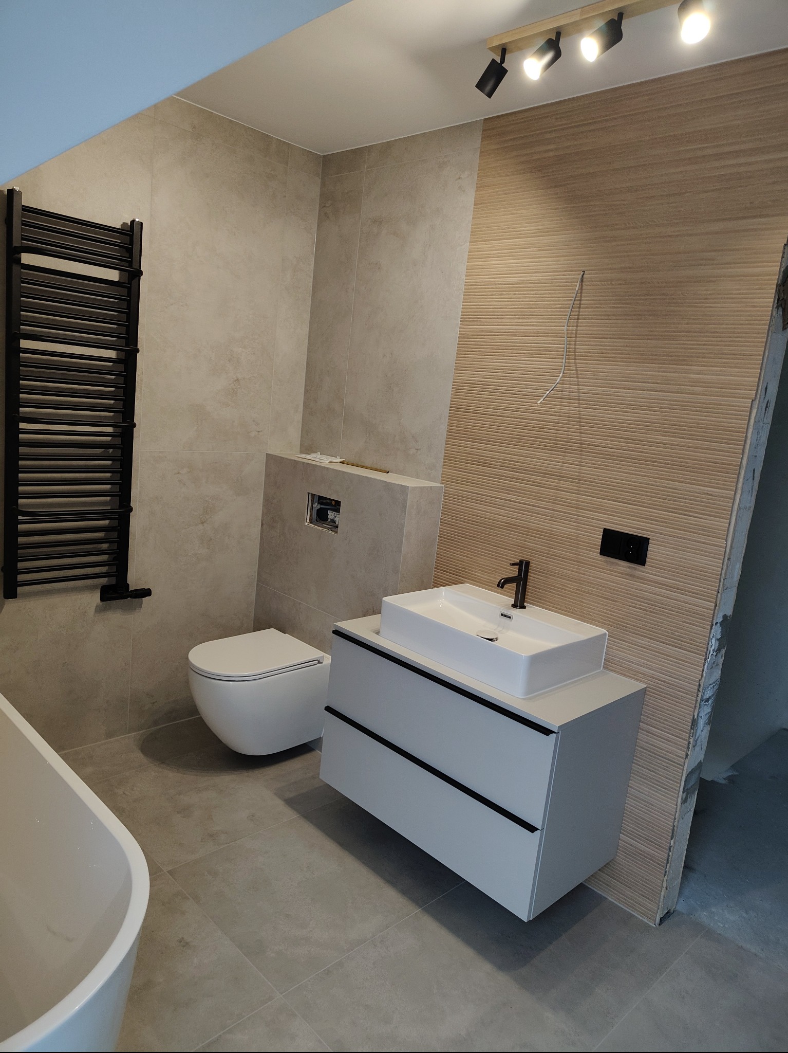 Nowoczesna łazienka po remoncie: beżowe płytki, biała umywalka z szafką, toaleta podwieszana i czarny grzejnik drabinkowy. Minimalistyczny design i ciepłe oświetlenie.