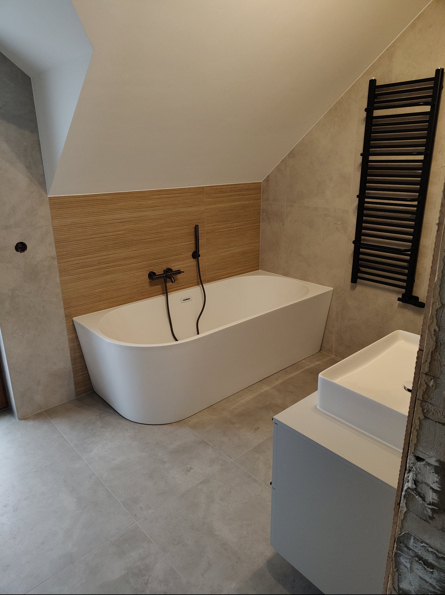 Nowoczesna łazienka na poddaszu z białą wanną, umywalką i czarnym grzejnikiem drabinkowym. Jasne płytki i drewnopodobne panele tworzą spójną całość.