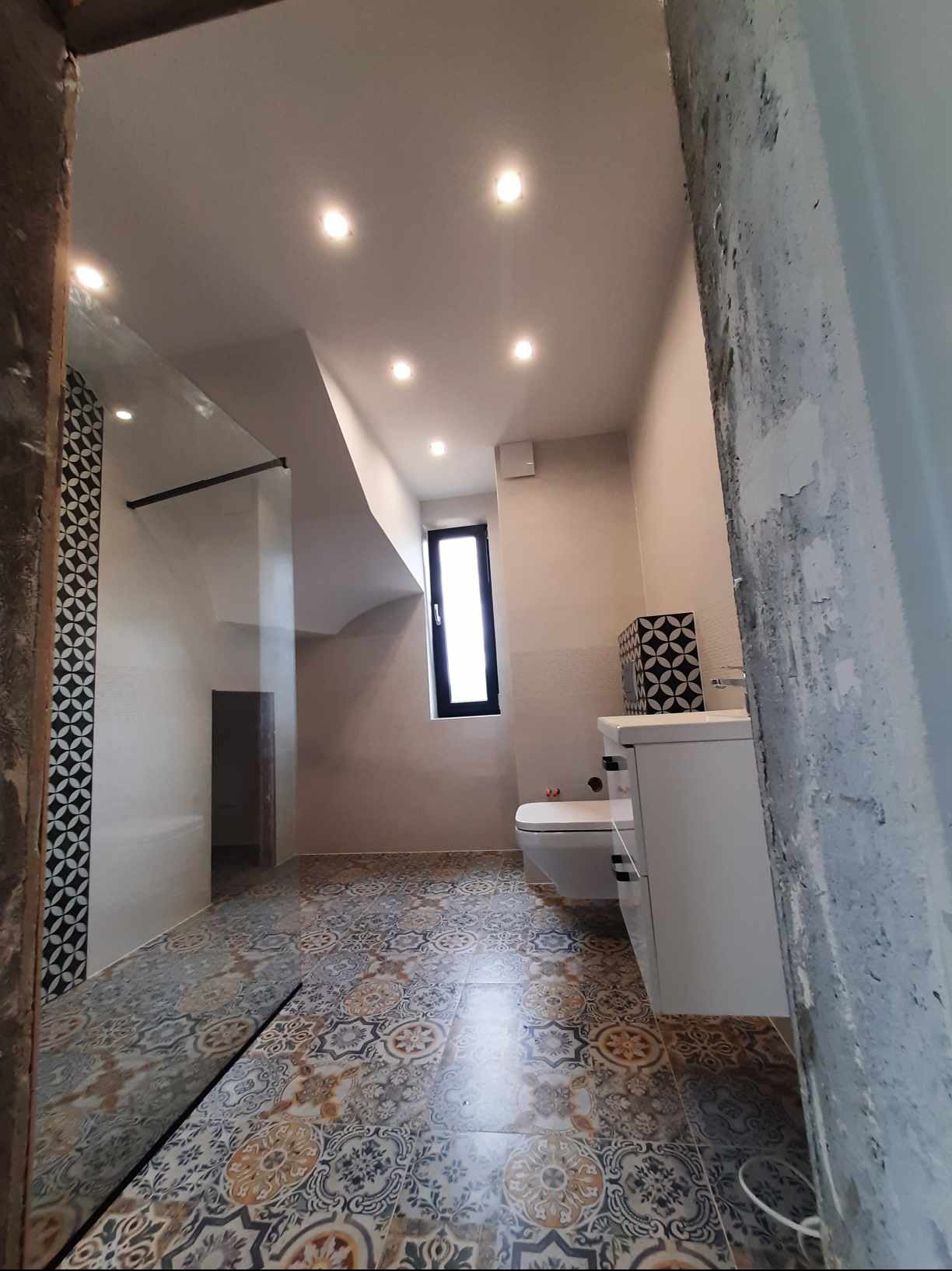 Wykończona łazienka z wzorzystą podłogą i kabiną prysznicową typu walk-in, widok zza surowej ściany w trakcie remontu. Nowoczesne oświetlenie punktowe.