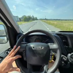 Widok z kabiny Iveco na drogę przez szybę z niewielkimi zabrudzeniami, z ręką kierowcy na kierownicy i widoczną deską rozdzielczą. Krajobraz wiejski w tle.