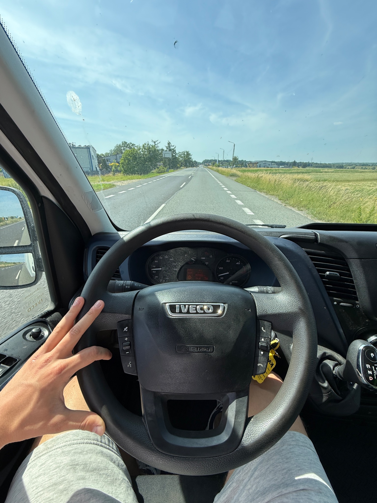 Widok z kabiny Iveco na drogę przez szybę z niewielkimi zabrudzeniami, z ręką kierowcy na kierownicy i widoczną deską rozdzielczą. Krajobraz wiejski w tle.