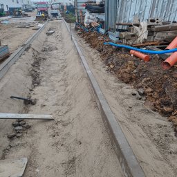 PRIME ROAD WIESŁAW BŁASIK - Przygotowanie podłoża: koryto wyłożone piaskiem, obrzeża betonowe, narzędzia pracy w tle. Etap prac ziemnych przed ułożeniem kostki. Widoczne rury i materiały budowlane.