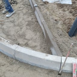 PRIME ROAD WIESŁAW BŁASIK - Układanie obrzeży betonowych na podsypce piaskowej. Widoczne narzędzia i fragment pracownika. Obrzeże łukowe i proste. Prace brukarskie w toku.
