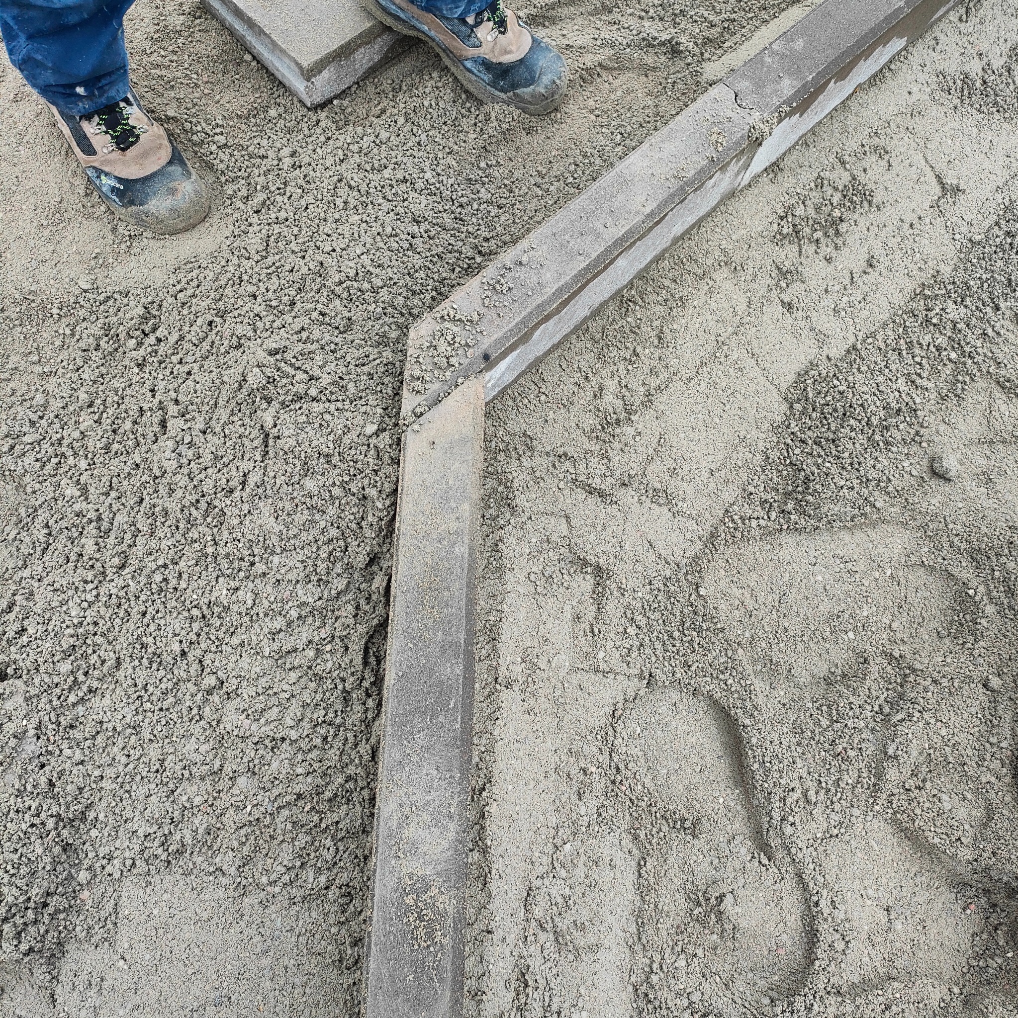 Etap przygotowania podłoża: obrzeża z betonu, podsypka piaskowa, robocze buty z widocznymi zabrudzeniami, kostka brukowa w tle. Układanie obrzeży.