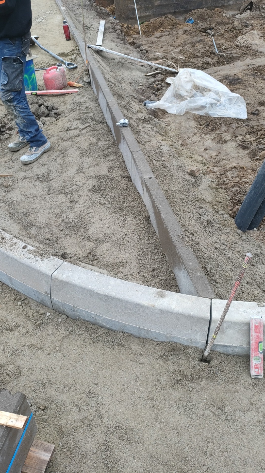 Układanie obrzeży betonowych na podsypce piaskowej. Widoczne narzędzia i fragment pracownika. Obrzeże łukowe i proste. Prace brukarskie w toku.