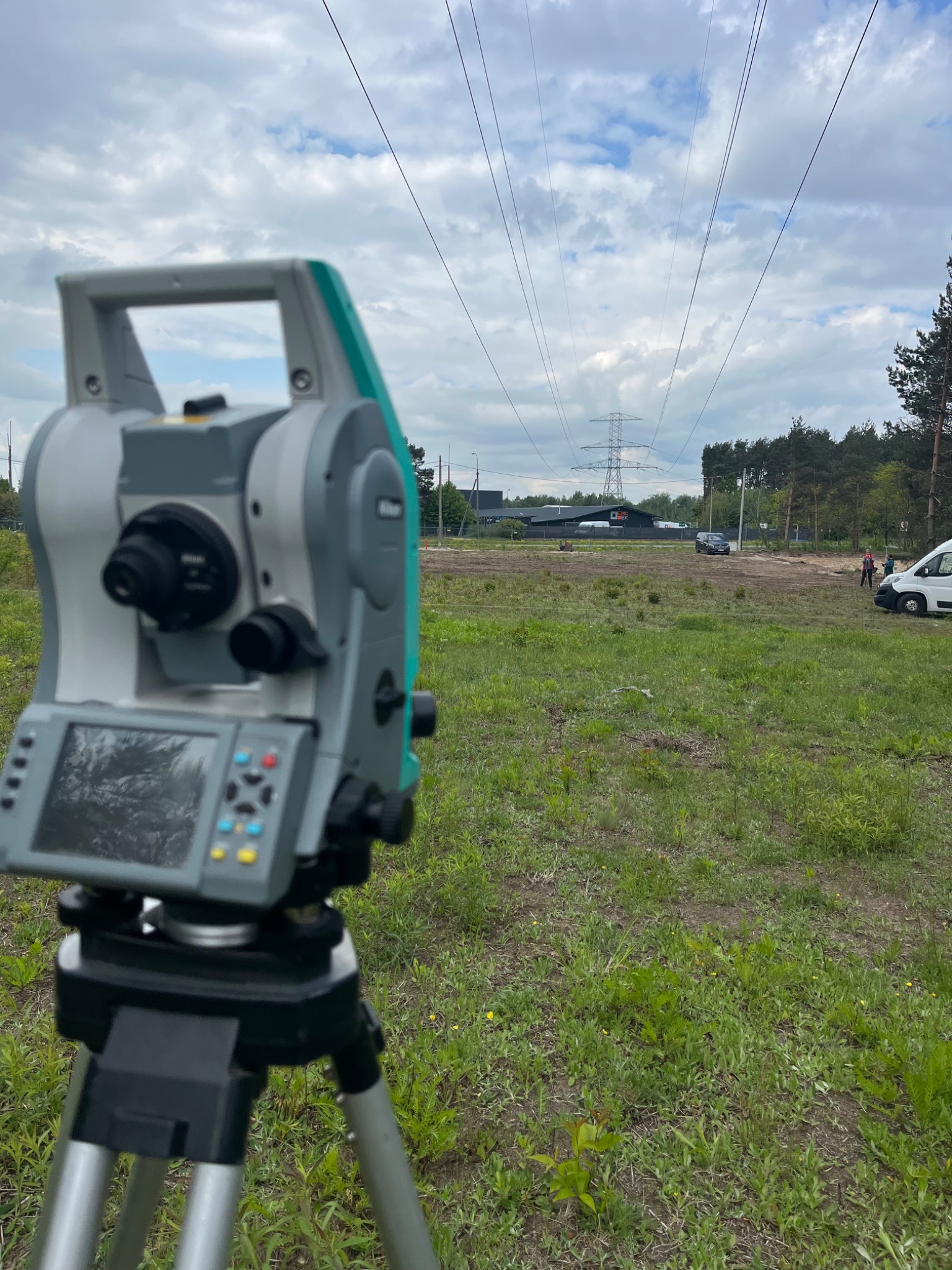 Geodezyjny tachimetr Nikon na trójnogu w terenie trawiastym, w tle słupy wysokiego napięcia i budynek. Prace geodezyjne w otwartym terenie.