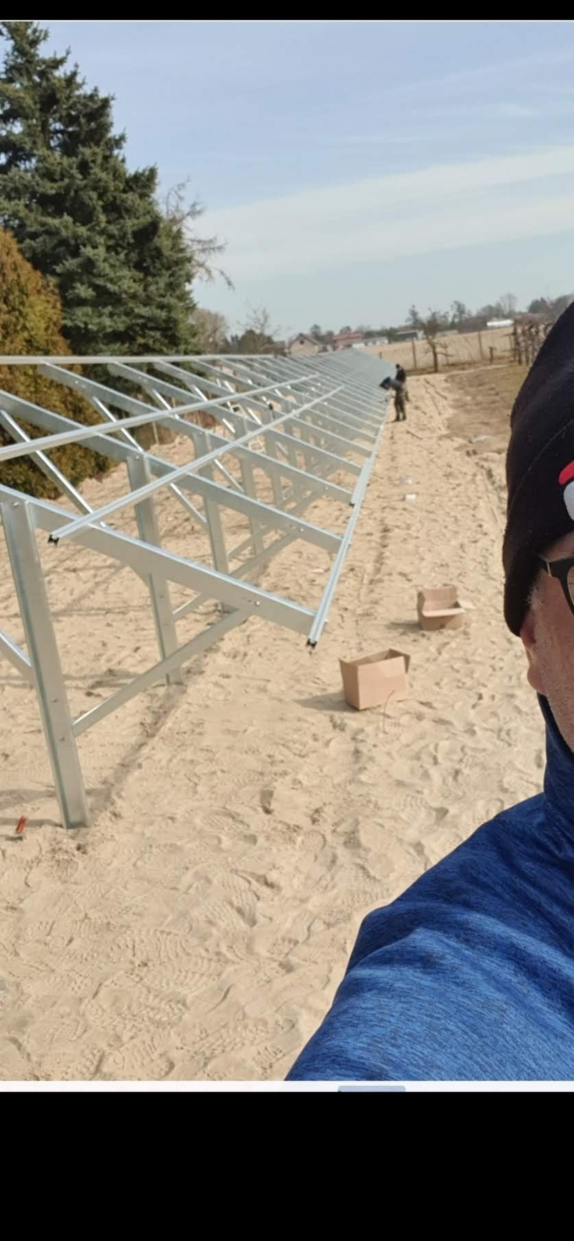 Konstrukcja szkieletowa przyszłej instalacji, prawdopodobnie fotowoltaicznej, z widocznymi elementami montażowymi i pracownikiem w oddali na piaszczystym terenie.