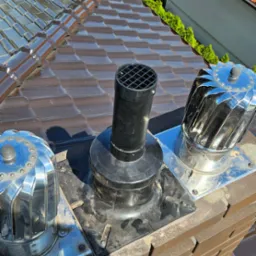 Widok z góry na komin z zainstalowanymi nasadami wentylacyjnymi i kominową, brązowa dachówka w tle.