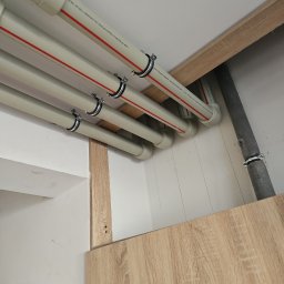 Przedsiębiorstwo Instalacyjne Kacper Kokociński - Białe rury z czerwonym paskiem, przymocowane metalowymi obejmami do drewnianej konstrukcji pod sufitem. Widoczna szara rura i instrukcja przeciwpożarowa.
