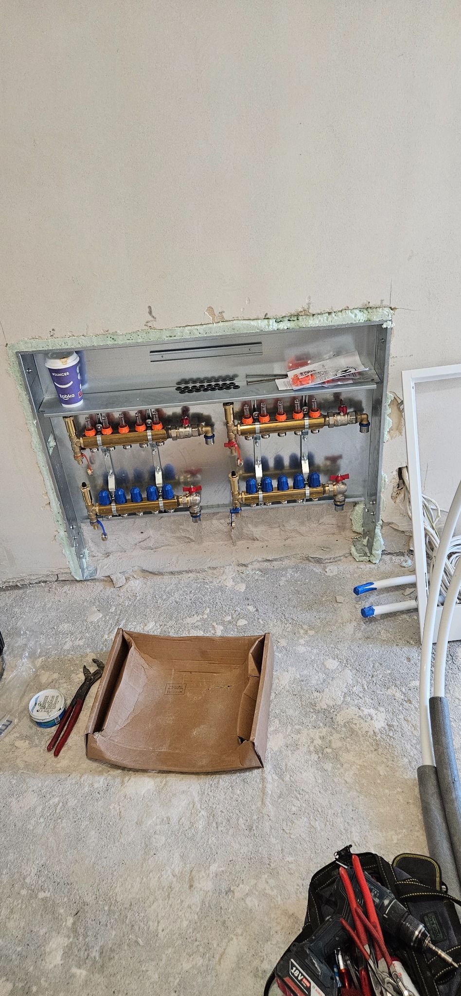 Rozdzielacz ogrzewania podłogowego w metalowej obudowie, zamontowany w ścianie. Widoczne narzędzia i akcesoria instalacyjne. Ściana i podłoga w stanie surowym.