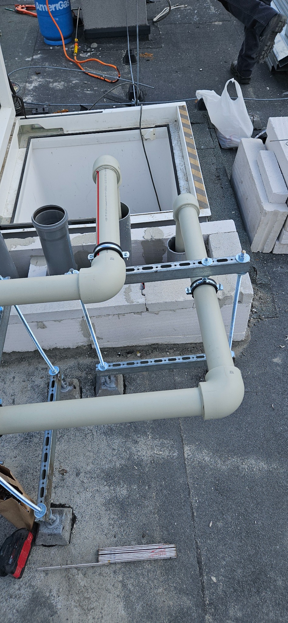 Instalacja rur wentylacyjnych na dachu budynku, wsparta na metalowych konstrukcjach. Widoczne elementy montażowe, bloczki betonowe i narzędzia.