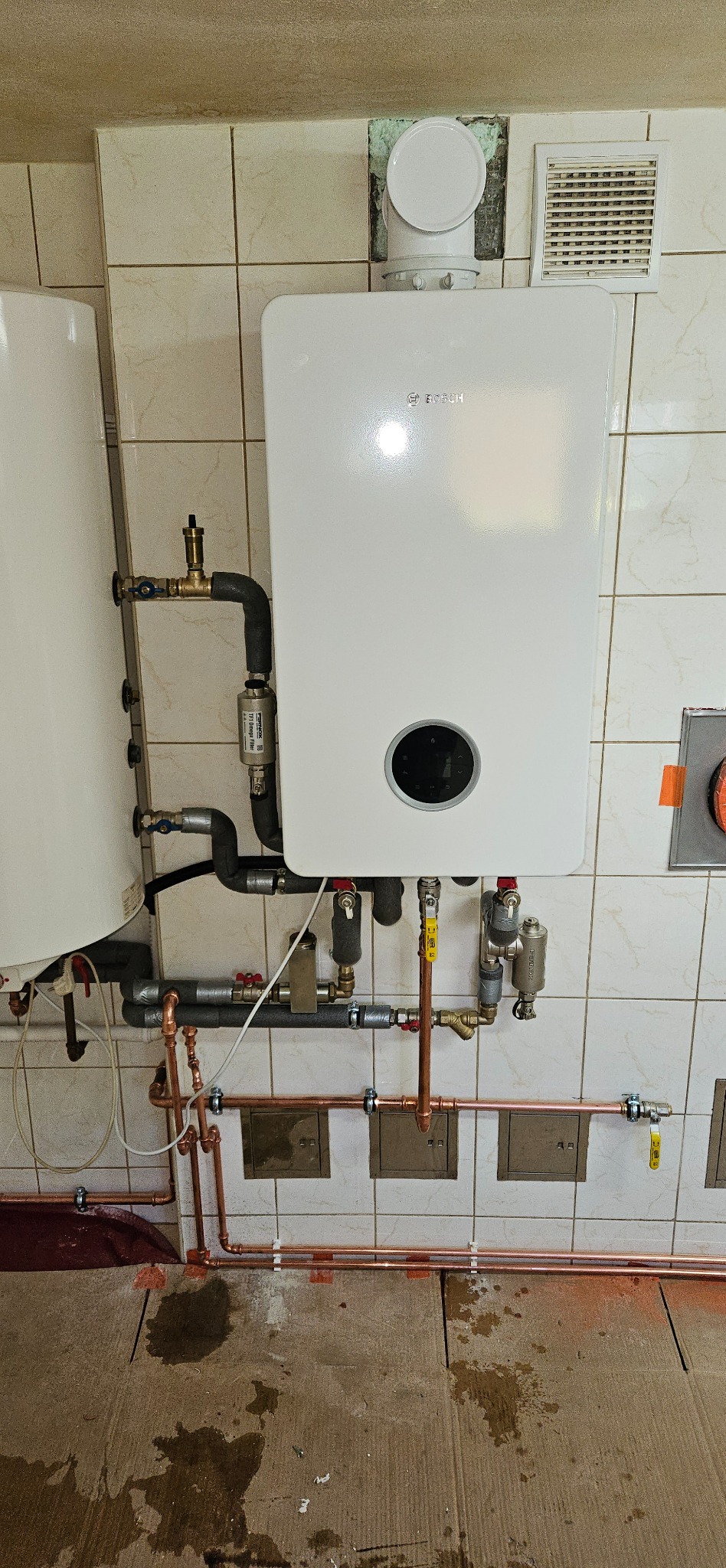 Nowoczesny, biały piec gazowy Bosch zamontowany na kafelkowanej ścianie, z widoczną instalacją hydrauliczną z miedzianych rur i zaworów. Detal montażu.