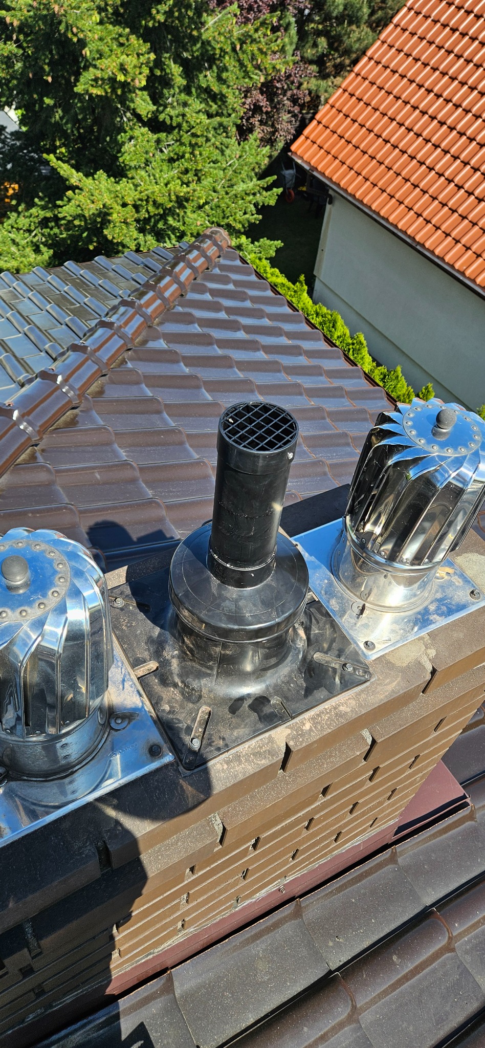 Widok z góry na komin z zainstalowanymi nasadami wentylacyjnymi i kominową, brązowa dachówka w tle.