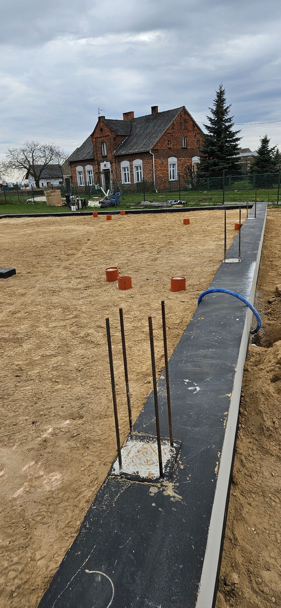 Fundament domu jednorodzinnego z wystającymi prętami zbrojeniowymi, widok z góry. W tle ceglany dom i drzewa.