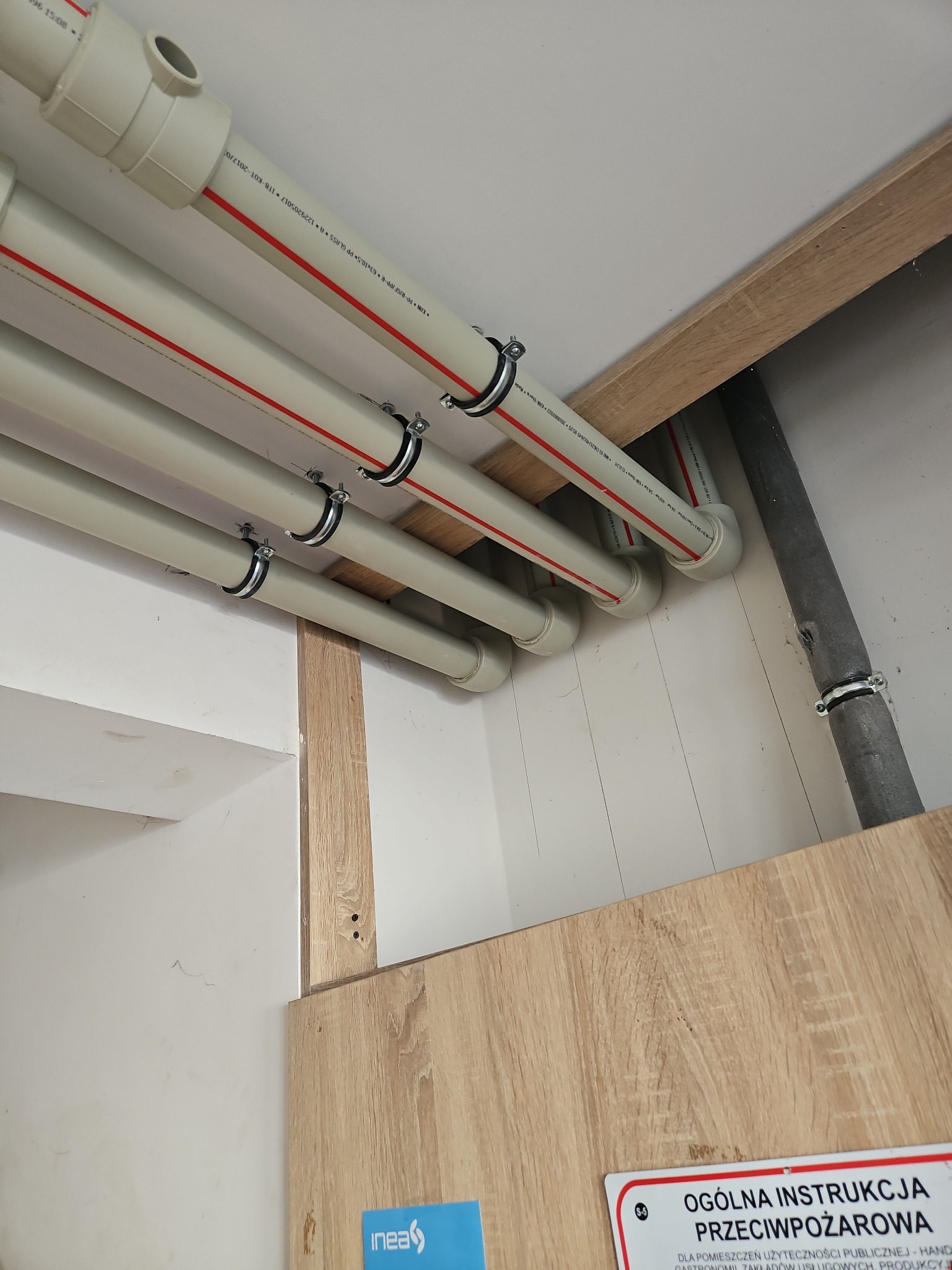 Białe rury z czerwonym paskiem, przymocowane metalowymi obejmami do drewnianej konstrukcji pod sufitem. Widoczna szara rura i instrukcja przeciwpożarowa.