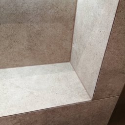 Patrykdys - Beżowa, teksturowana okładzina ceramiczna w narożniku wnęki. Precyzyjne łączenia płytek, widoczne delikatne żyłki i subtelne światło w górnej części wnęki.