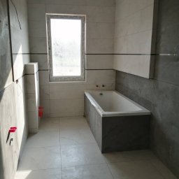 Patrykdys - Nowoczesna łazienka po remoncie: białe płytki imitujące marmur, szara zabudowa wanny, okno z widokiem na zewnątrz. Minimalistyczny design, świeżo po wykończeniu.