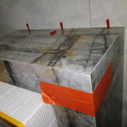 Patrykdys - Montaż płytek imitujących marmur na zabudowie, z użyciem klipsów poziomujących i taśmy zabezpieczającej. Widoczny fragment paczki z płytkami.