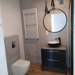 Patrykdys - Nowoczesna łazienka z szarą glazurą, wiszącą toaletą, grzejnikiem drabinkowym i okrągłym lustrem nad czarną szafką z umywalką.