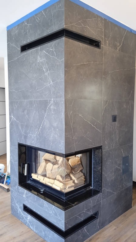 Obudowa kominkowa z szarych płyt imitujących marmur, z czarnymi wstawkami i drewnem w palenisku. Widoczny fragment podłogi i akcesoria malarskie w tle.