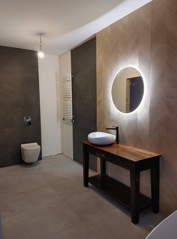Nowoczesna łazienka z podświetlanym lustrem, drewnianą konsolą pod umywalką i płytkami w odcieniach szarości i beżu. Minimalistyczny design.