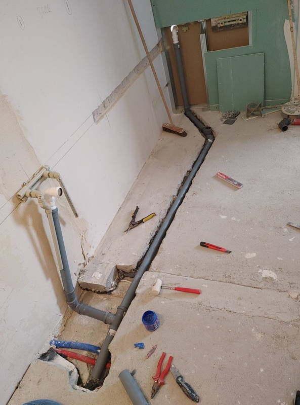 Montaż szarej instalacji kanalizacyjnej w wyżłobionej bruździe w podłodze. Wokół widoczne narzędzia i akcesoria instalacyjne. Częściowy widok pomieszczenia w trakcie remontu.