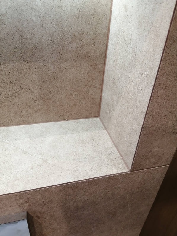 Beżowa, teksturowana okładzina ceramiczna w narożniku wnęki. Precyzyjne łączenia płytek, widoczne delikatne żyłki i subtelne światło w górnej części wnęki.