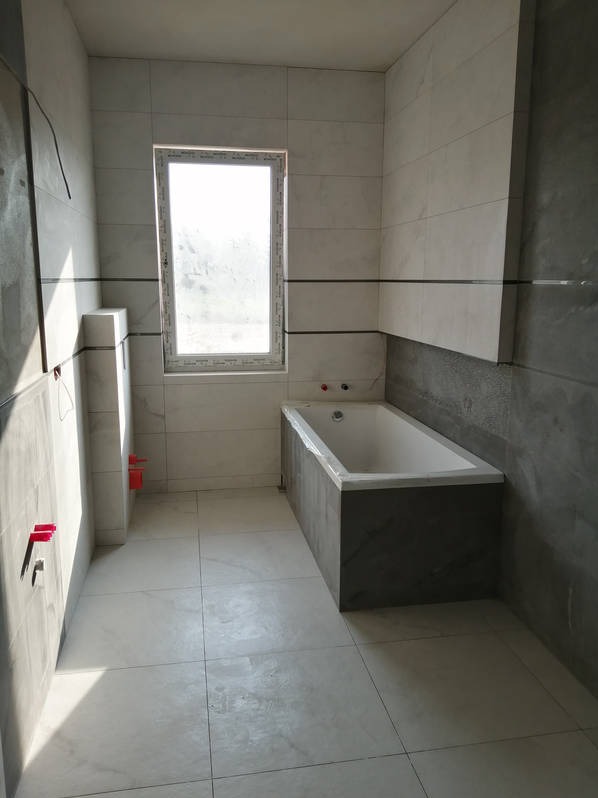 Nowoczesna łazienka po remoncie: białe płytki imitujące marmur, szara zabudowa wanny, okno z widokiem na zewnątrz. Minimalistyczny design, świeżo po wykończeniu.