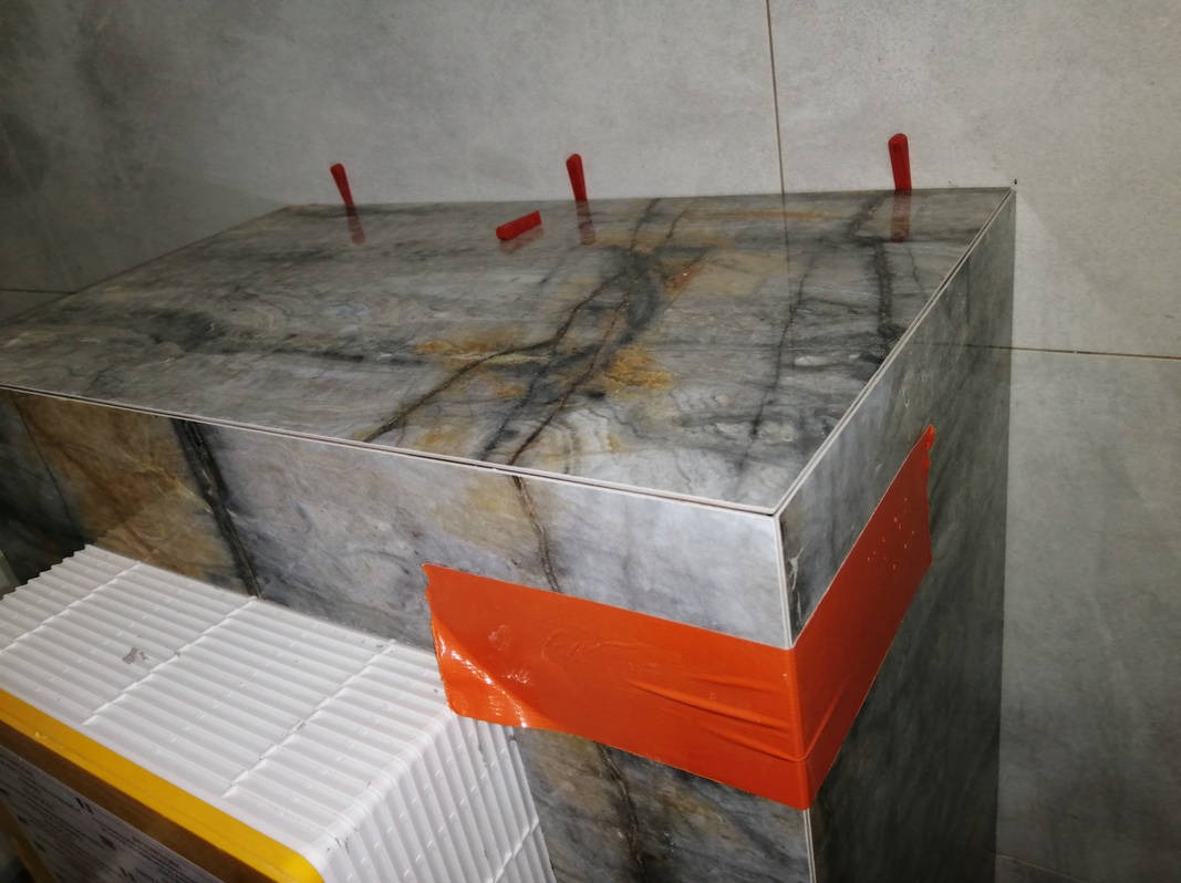 Montaż płytek imitujących marmur na zabudowie, z użyciem klipsów poziomujących i taśmy zabezpieczającej. Widoczny fragment paczki z płytkami.