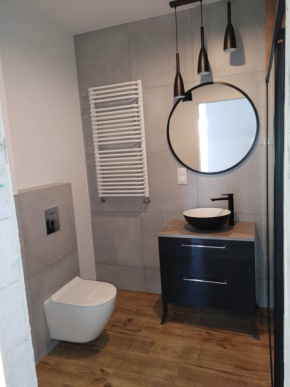Nowoczesna łazienka z szarą glazurą, wiszącą toaletą, grzejnikiem drabinkowym i okrągłym lustrem nad czarną szafką z umywalką.