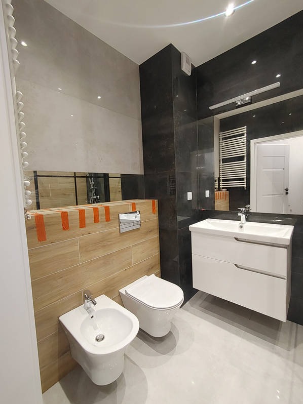 Nowoczesna łazienka z wiszącą toaletą, bidetem i umywalką z szafką. Na ścianach płytki imitujące drewno i ciemny kamień. Lustro na ścianie powiększa przestrzeń.