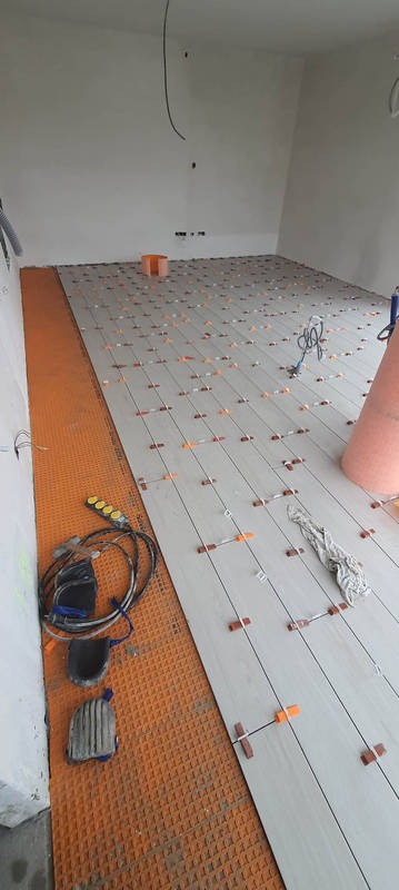 Układanie płytek podłogowych w jasnym pomieszczeniu, widoczne krzyżyki dystansowe i mata izolacyjna. Przewody i narzędzia glazurnicze na podłodze.