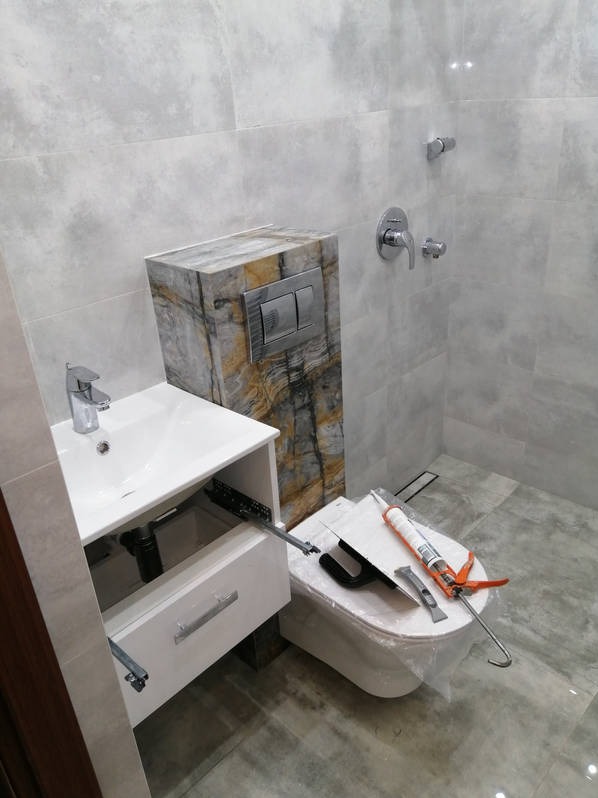 Montaż umywalki z szafką i podwieszanej toalety w łazience z szarymi płytkami. Widoczne narzędzia i materiały montażowe.