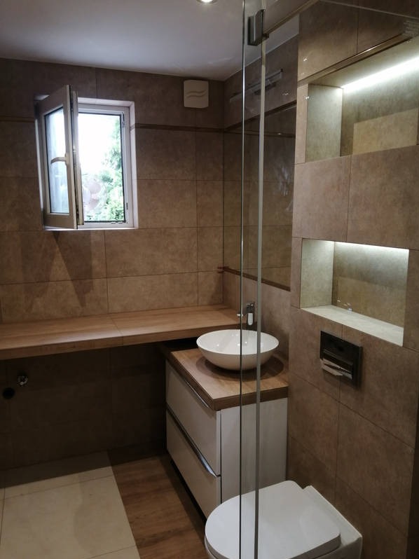 Jasna łazienka z beżowymi płytkami, drewnianym blatem i białą umywalką. Kabina prysznicowa ze szklanymi drzwiami. Otwierane okno wpuszcza naturalne światło.