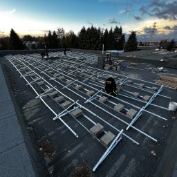 CENTRUM REALIZACJI PROSTA SPÓŁKA AKCYJNA - Montaż konstrukcji wsporczej pod panele słoneczne na dachu budynku. Widoczne metalowe profile, betonowe bloczki i pracownik w stroju roboczym. Tło: drzewa i zachodzące słońce.