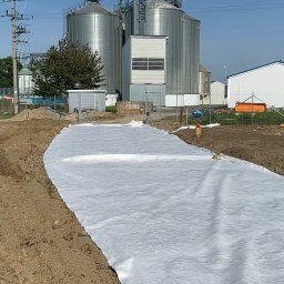 CENTRUM REALIZACJI PROSTA SPÓŁKA AKCYJNA - Przygotowanie terenu pod instalację: rozłożona biała geowłóknina na wykopie, w tle silosy zbożowe i budynek z turbinami wiatrowymi.