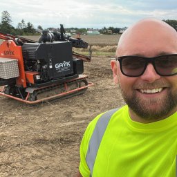 CENTRUM REALIZACJI PROSTA SPÓŁKA AKCYJNA - Mężczyzna w okularach i jaskrawej koszulce pozuje na tle pomarańczowej wiertnicy geologicznej na gąsienicach, stojącej na polu z niską roślinnością.