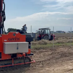 Montaż słupków w polu z użyciem specjalistycznej maszyny GAYK i traktora. Widoczne ustawione słupki, pole po żniwach i domy w oddali. Praca w toku.