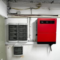 CENTRUM REALIZACJI PROSTA SPÓŁKA AKCYJNA - Czerwony inwerter GoodWe, dwie szare skrzynki elektryczne i mniejsza skrzynka na ścianie w pomieszczeniu. Widoczne kable i metalowe korytka kablowe.